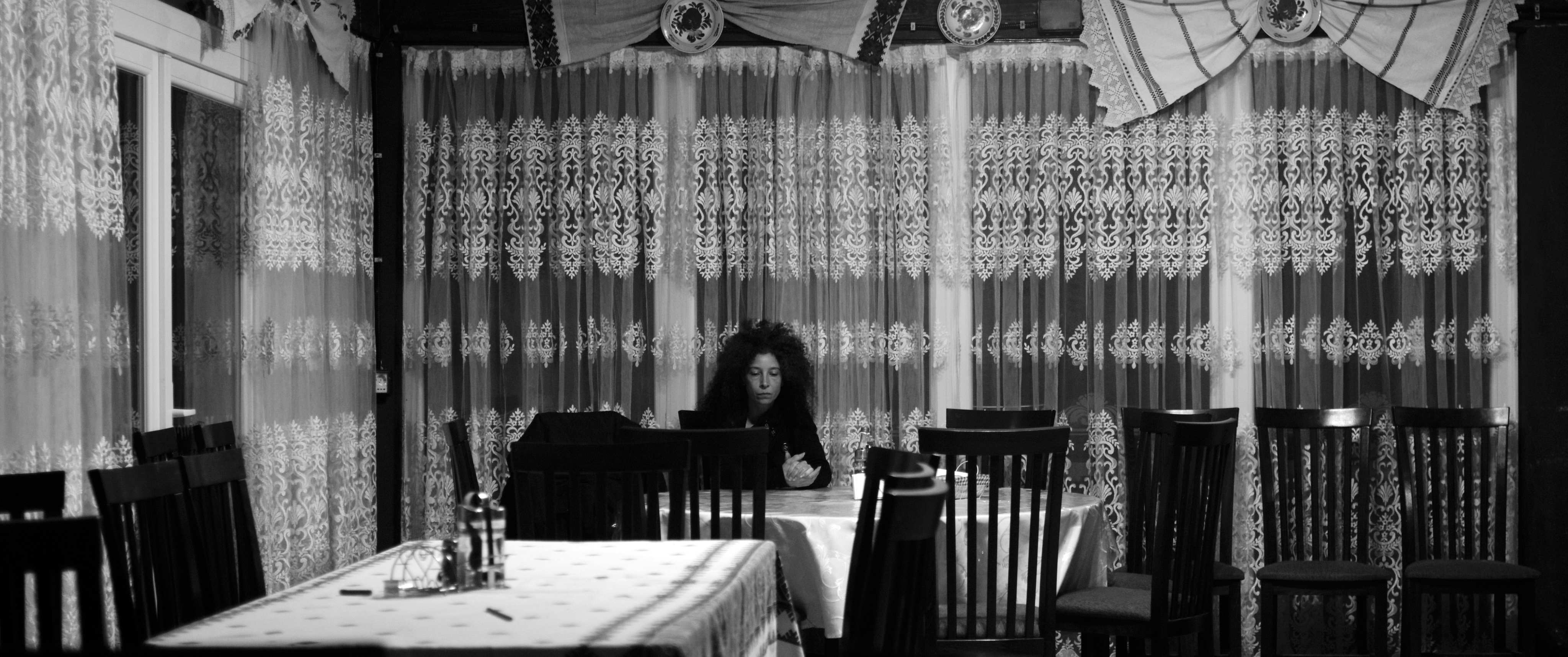Black and white frontal shot of Gheorghe looking into the camera. He is sitting at a table in the courtyard of his house. In the background, we see the house and a swingset. To the right of the picture and above, there is a vineyard.