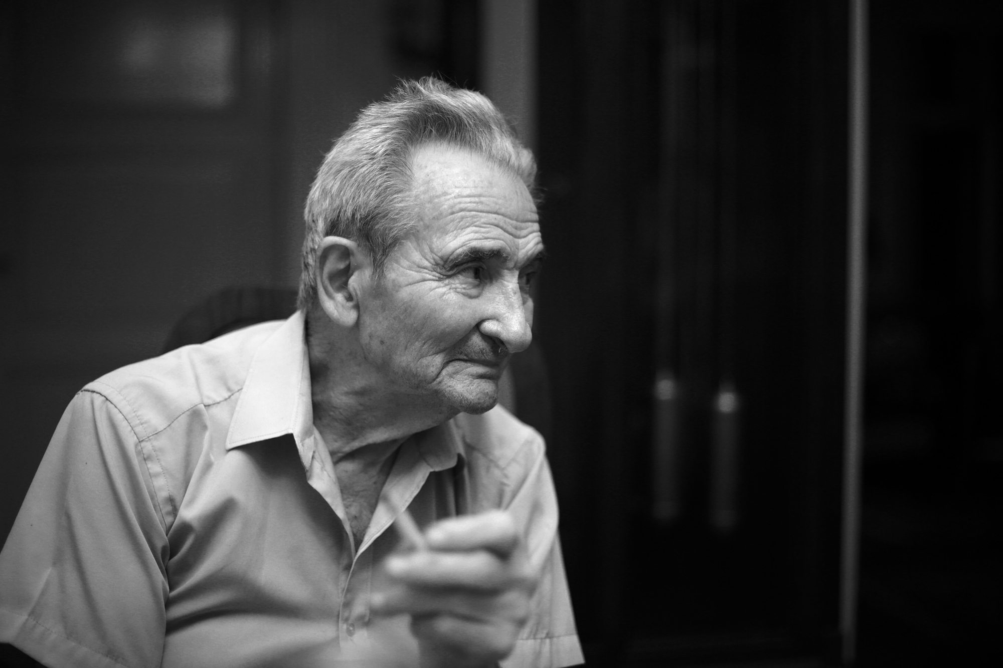 Black and white photograph of Licu looking to the right of the image. He is holding a cigarette. In the background, slightly out of focus, the pendulum of a clock.