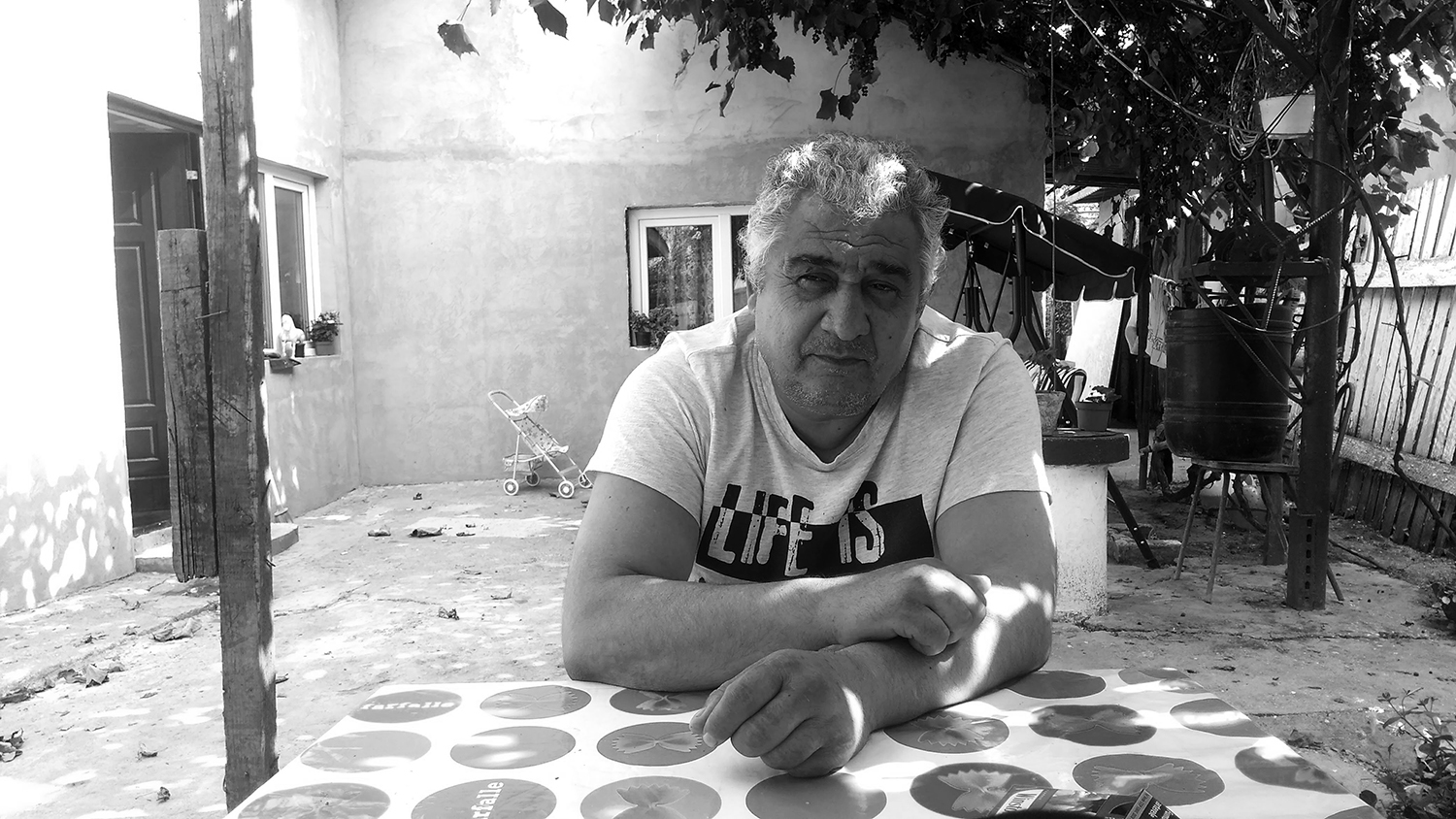 Black and white frontal shot of Gheorghe looking into the camera. He is sitting at a table in the courtyard of his house. In the background, we see the house and a swingset. To the right of the picture and above, there is a vineyard.
