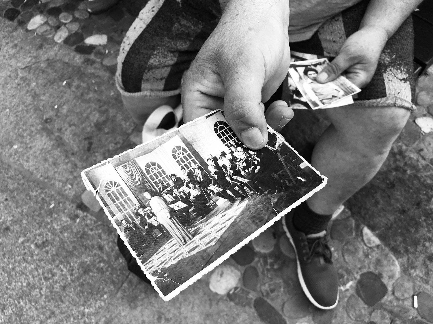 Close-up black and white photograph of a photograph held out by Gheorghe's right hand. It shows an orchestra and a singer at the time of communism. The left hand is holding other photographs. 
