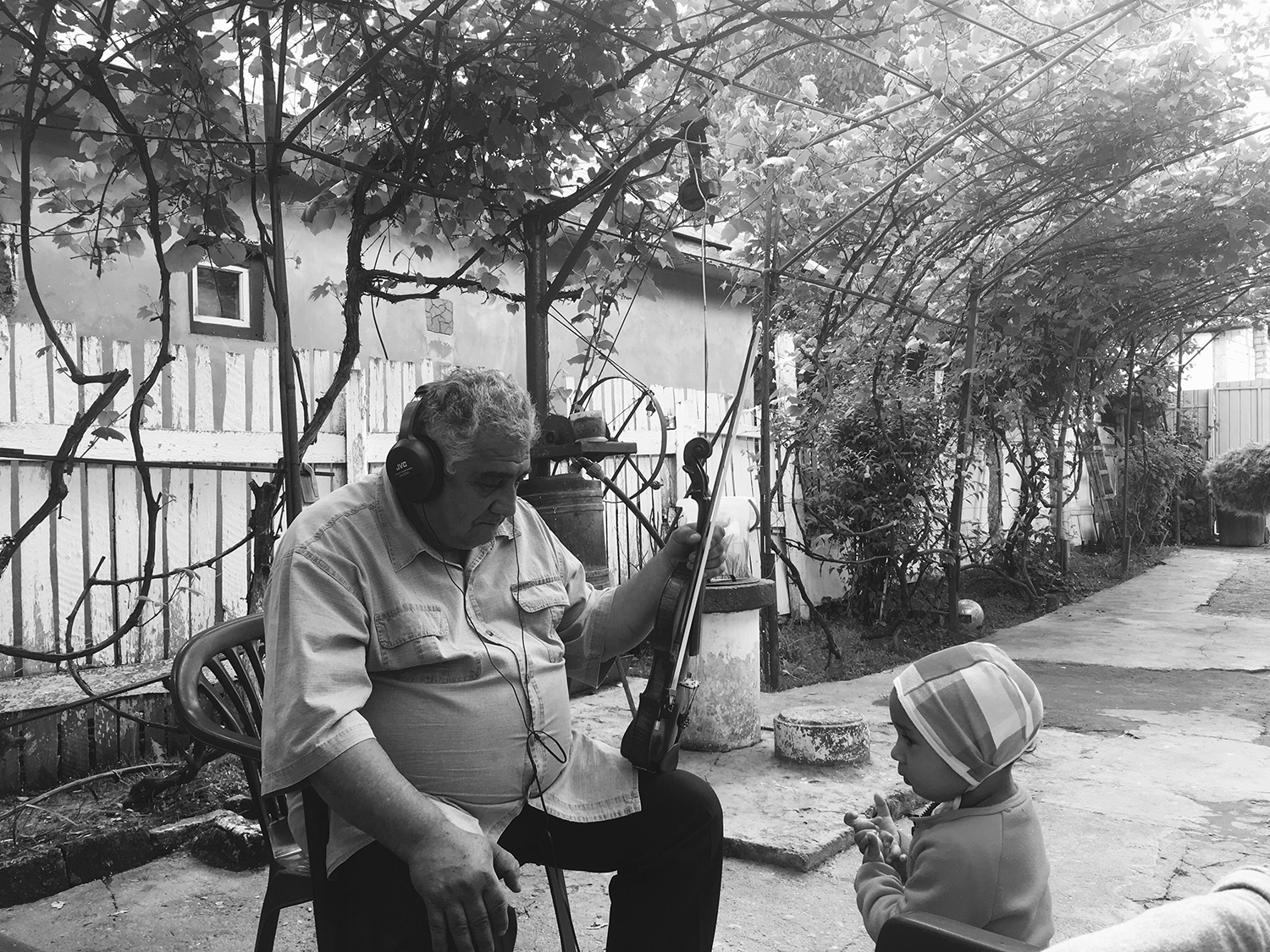 Black and white photograph of Gheorghe sitting on a garden chair in the courtyard of his house. His left hand is holding his violin and archer. His right hand is on his knee. He is listening attentively through headphones that he wears over his ears. Opposite him is a young child.